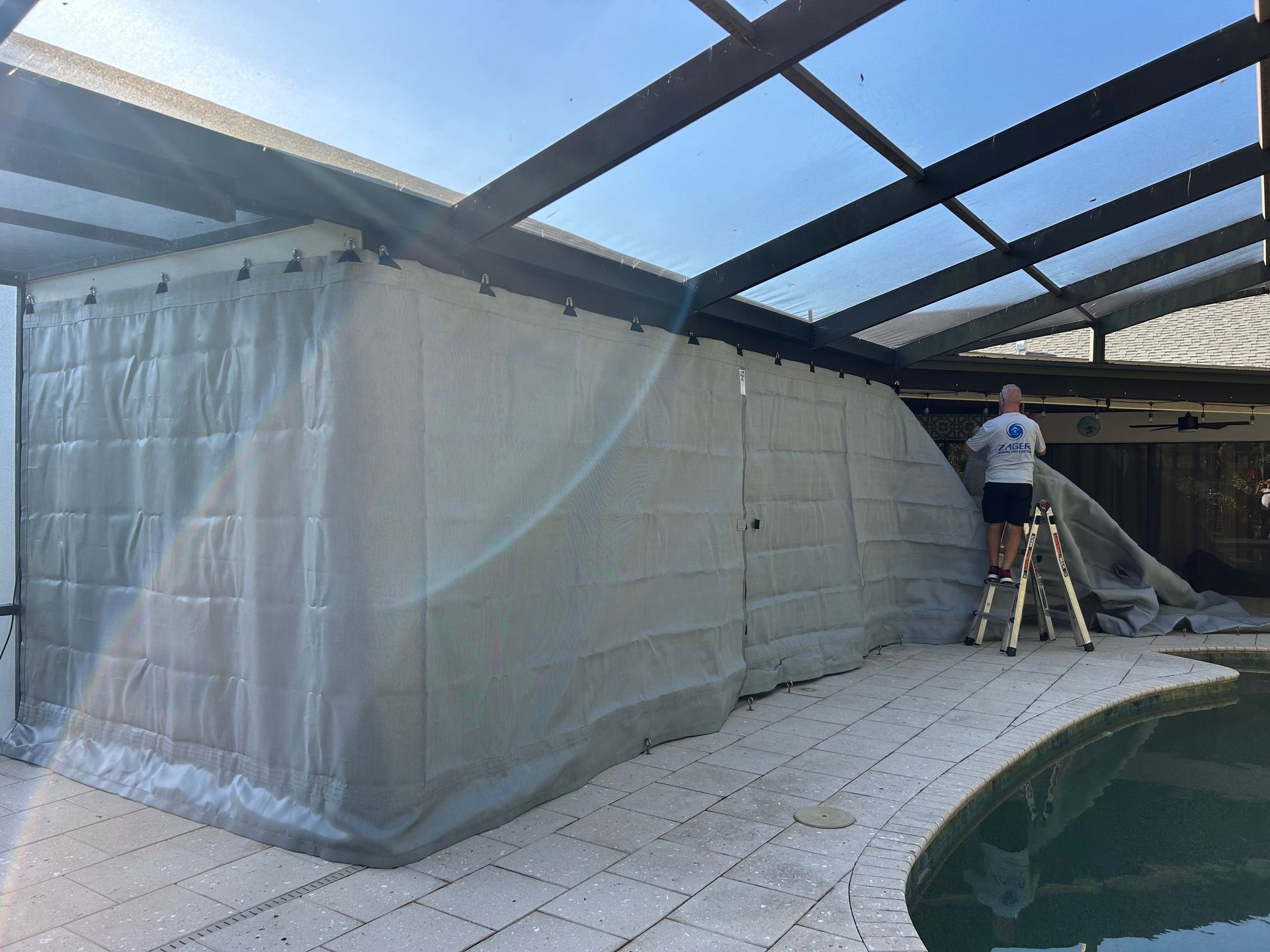 Man on a ladder installing a light gray curtain around a patio enclosure near a pool.