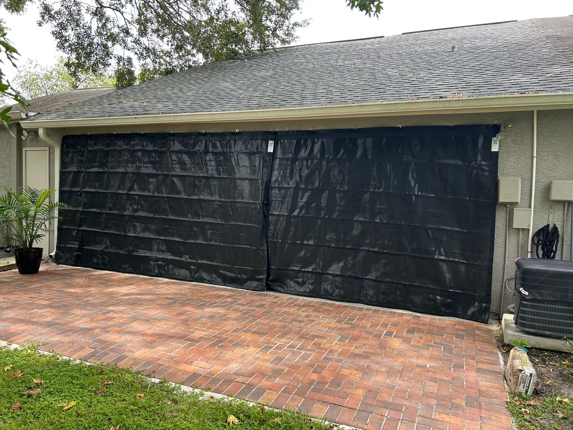 Black tarp covers a garage door on a brick patio. A potted plant sits to the left; an AC unit is to the right.