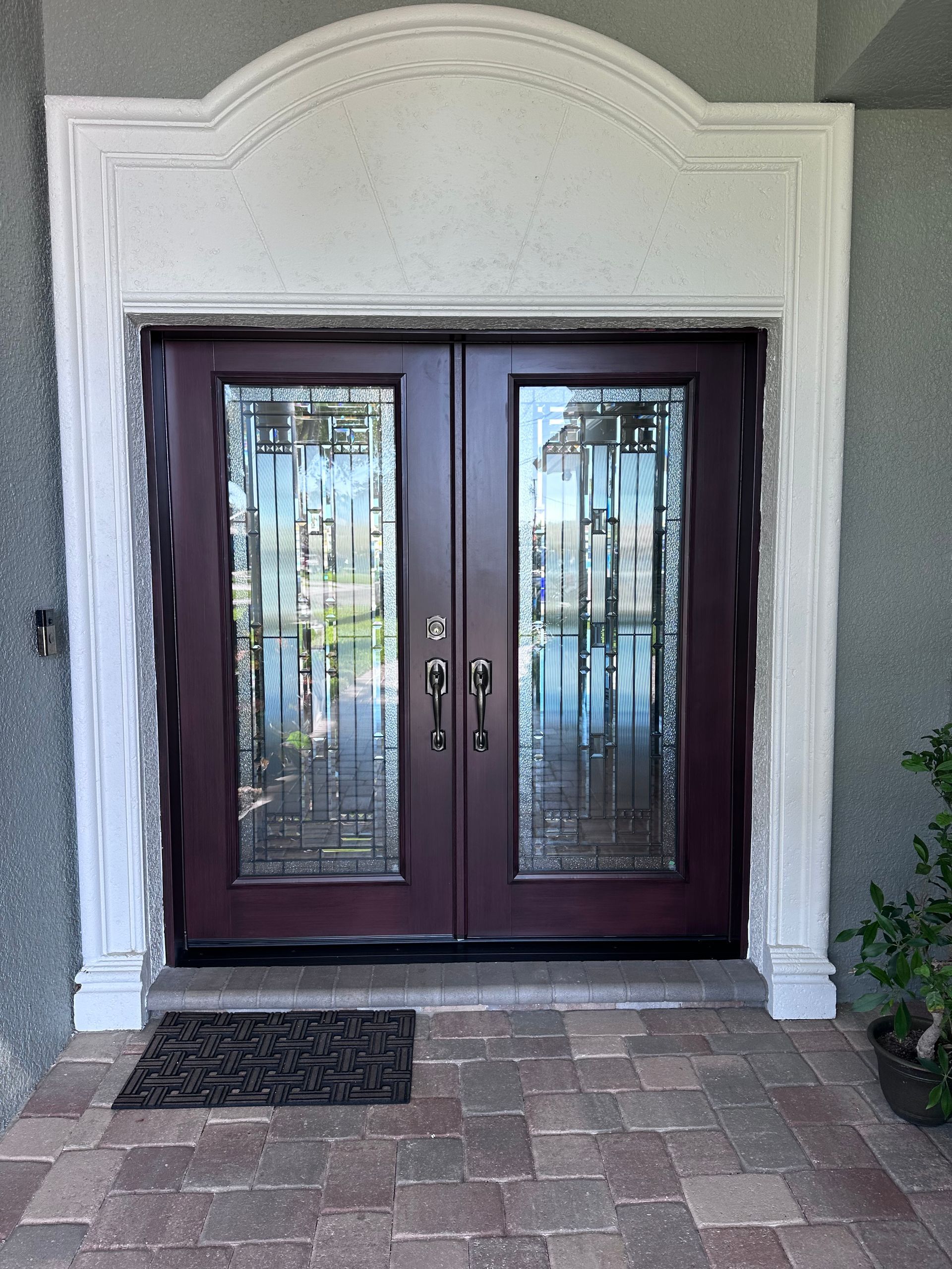 Double burgundy front doors with decorative glass panels, framed by white molding, dark welcome mat on brick walkway.