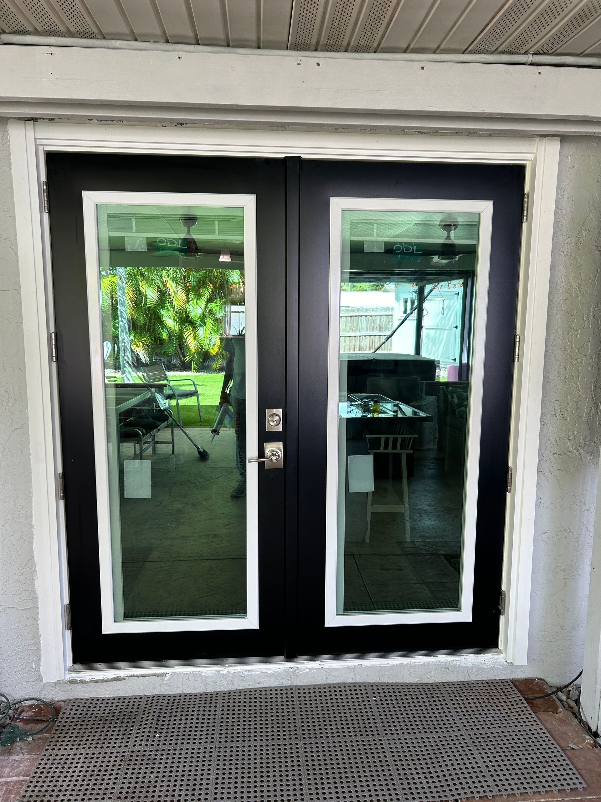 Black double doors with glass panels and white trim, set in a stucco frame, and a welcome mat.