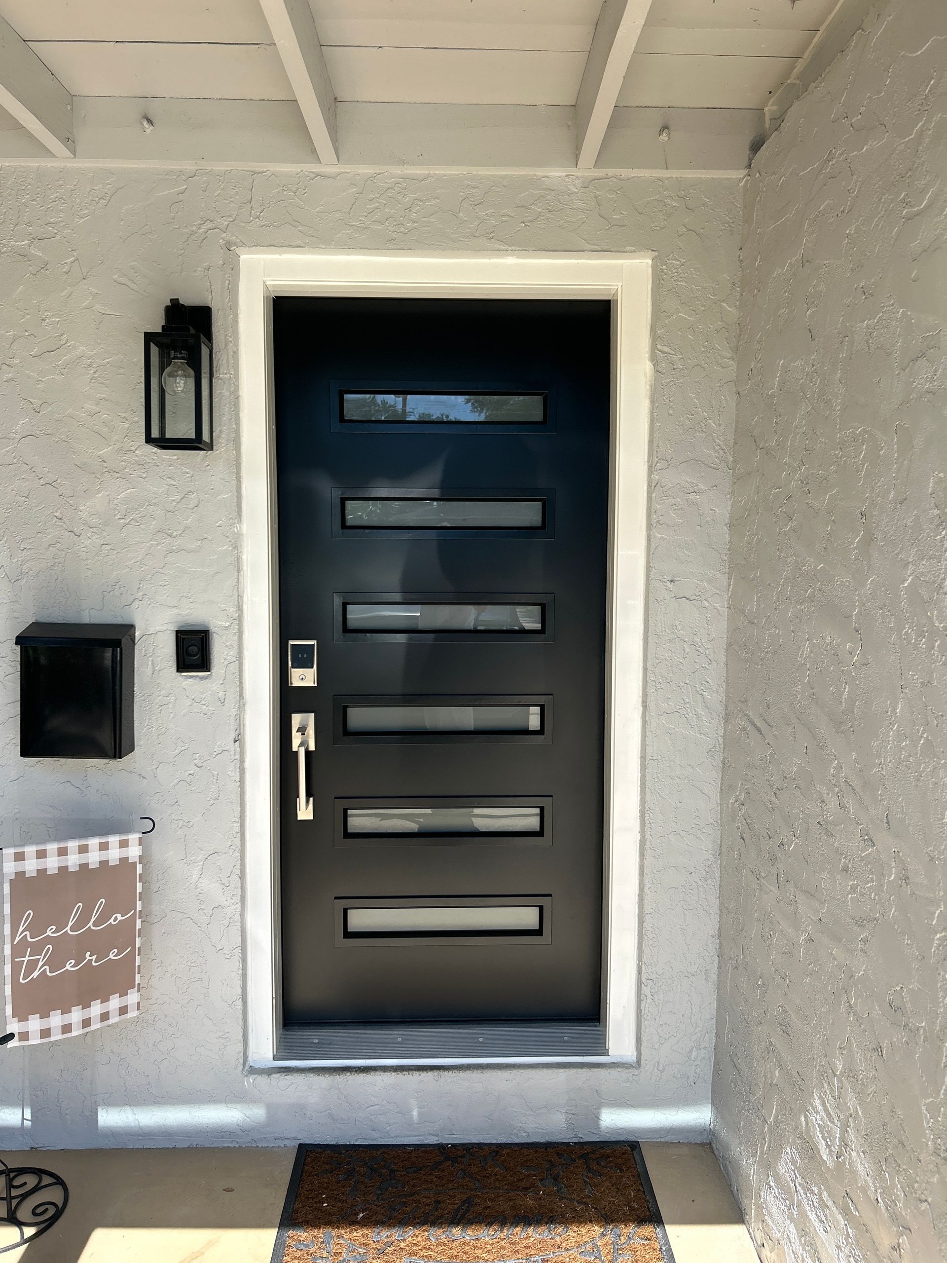 Black front door with horizontal glass panes and silver hardware set in a white-trimmed gray stucco wall.
