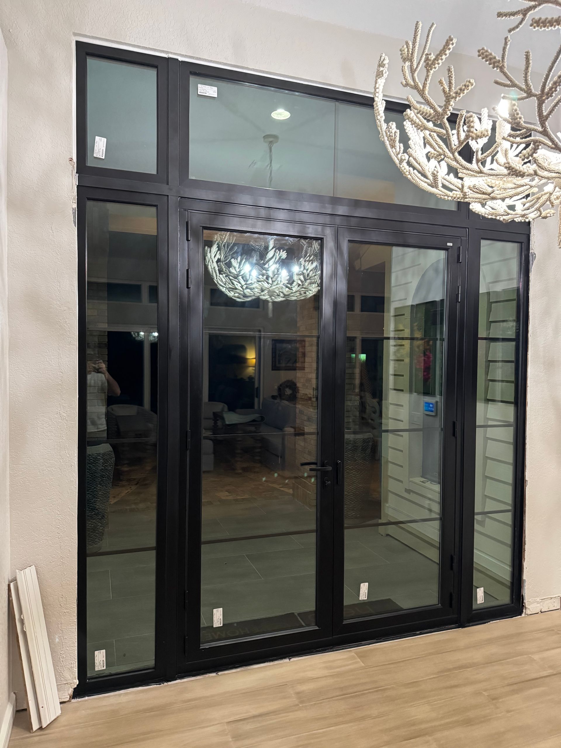 Black-framed glass doors and window. A modern interior shows a chandelier, light-colored walls, and wood floor.
