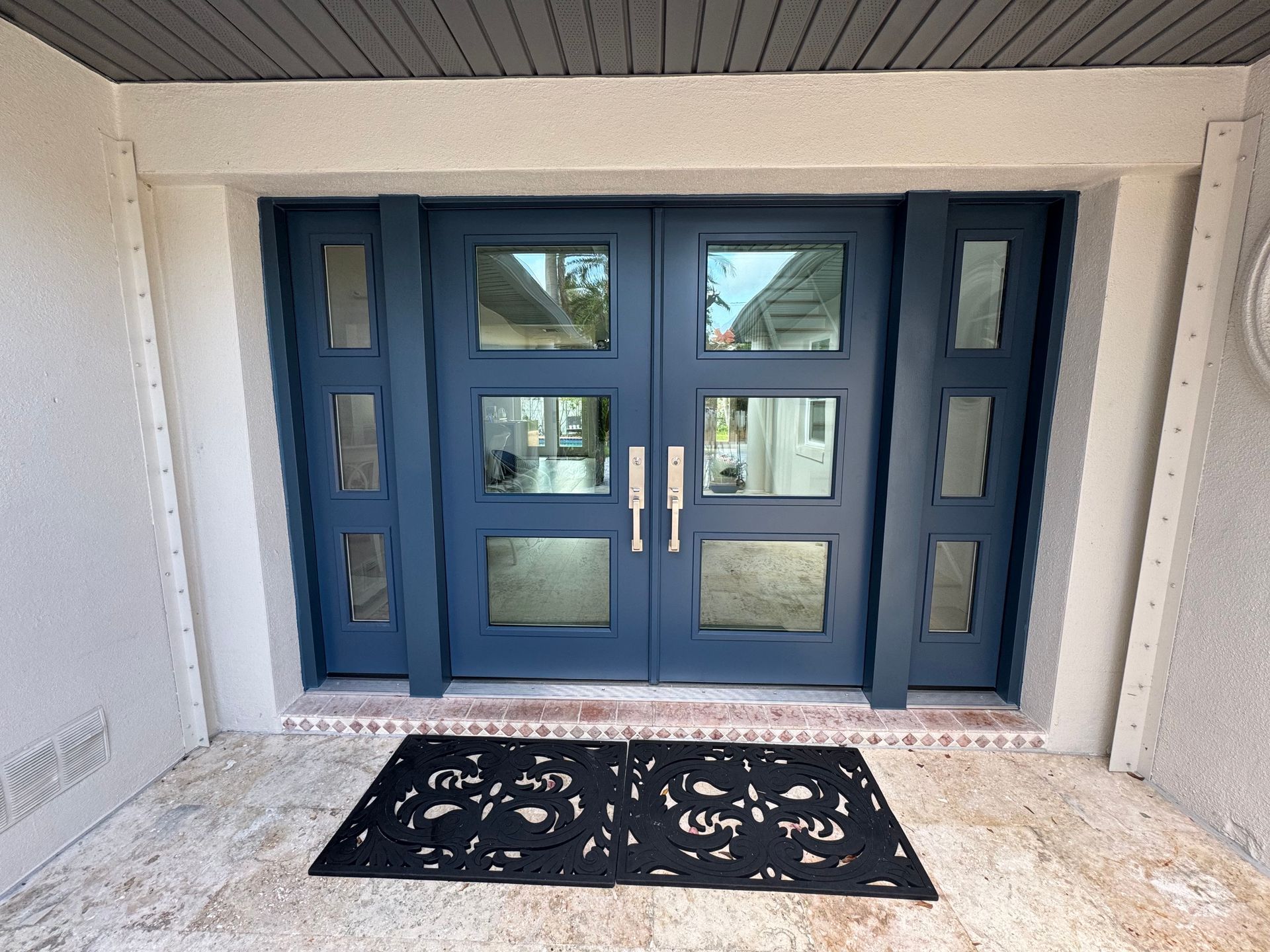 Blue double doors with glass panes, flanked by sidelights. Black doormat on light-colored stone patio.