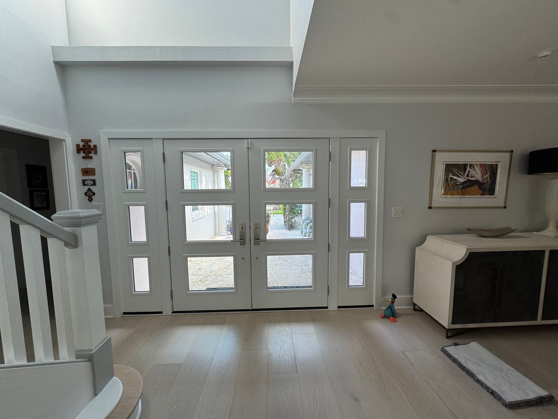 Bright entryway with white door and sidelights, light-colored wood floor, and a sideboard with a painting.