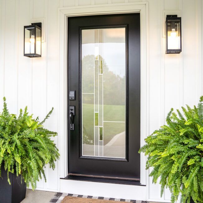 Black front door with glass panels, flanked by two outdoor lights and potted ferns on white siding.
