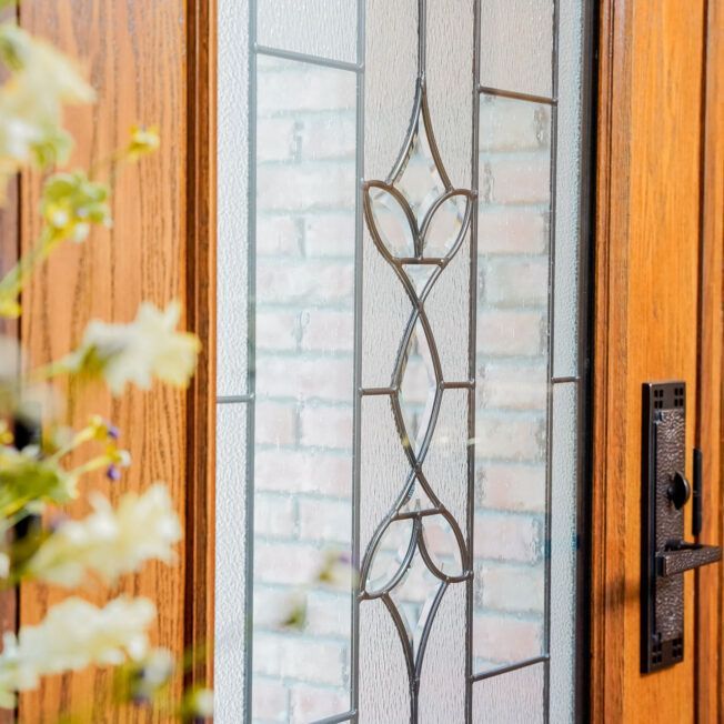 Wooden front door with a decorative glass panel, featuring a stylized floral design, with a brick wall visible behind the glass.