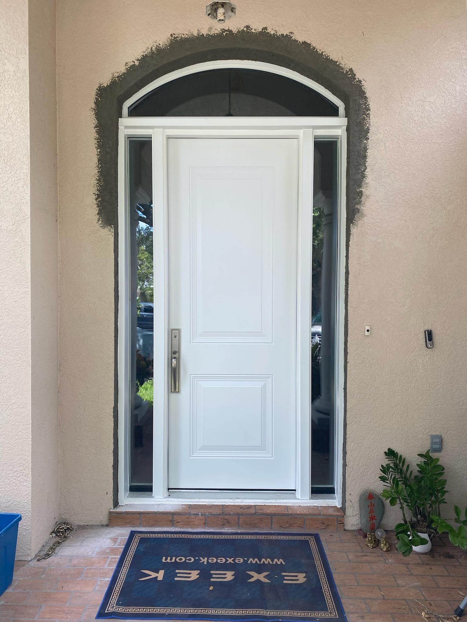 White front door with sidelights under an arched entryway, set in stucco siding. Blue welcome mat.