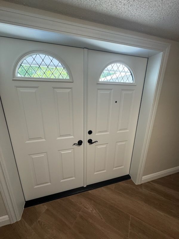 White double doors with arched, leaded glass panels, black hardware, and brown flooring.