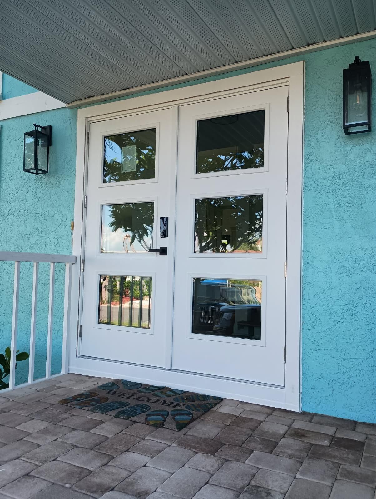 White double doors with glass panels on a blue stucco wall, black lanterns, and a welcome mat.