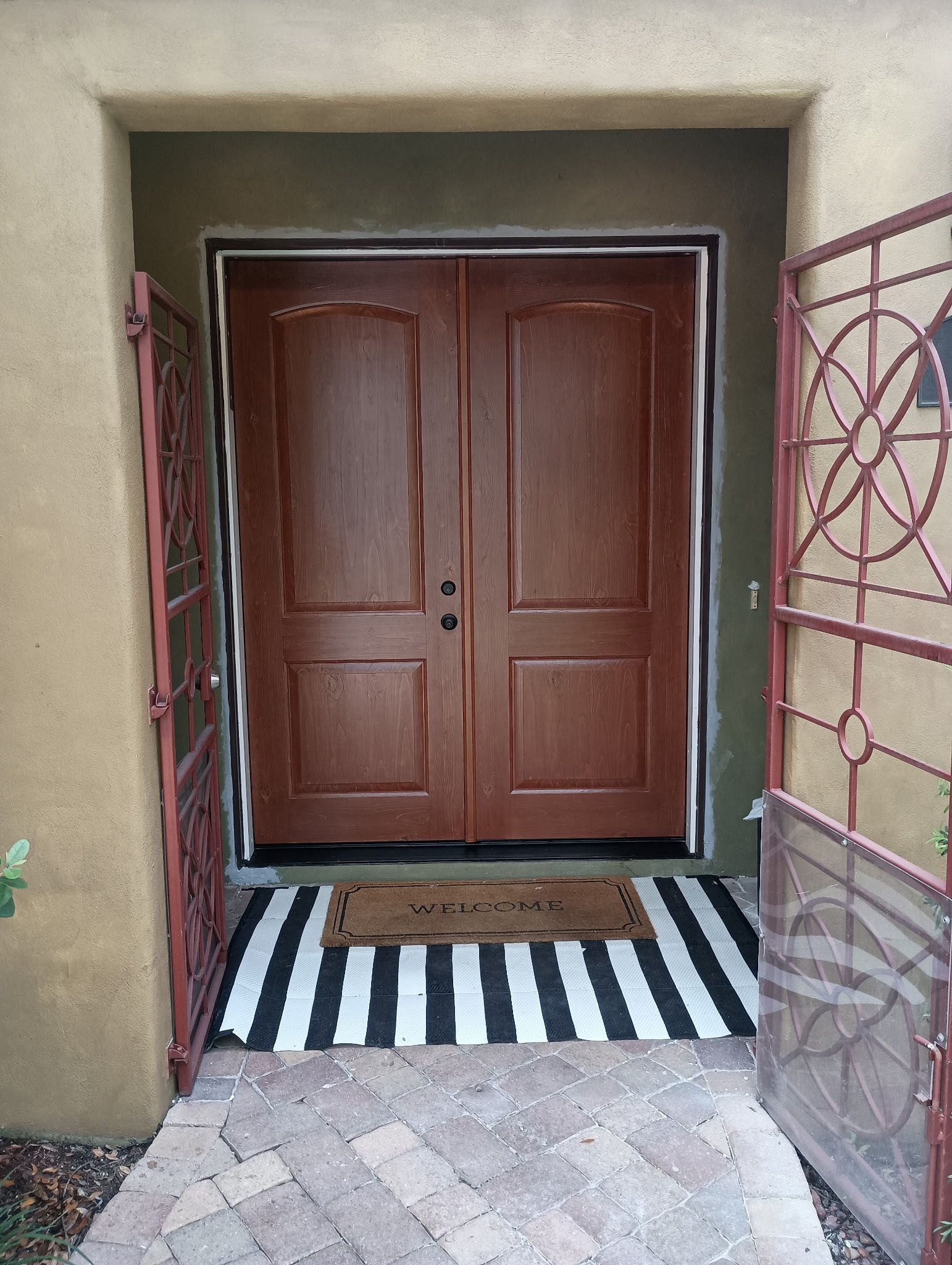 Exterior view of a brown double door with a welcome mat, black and white striped rug, and red security gates.