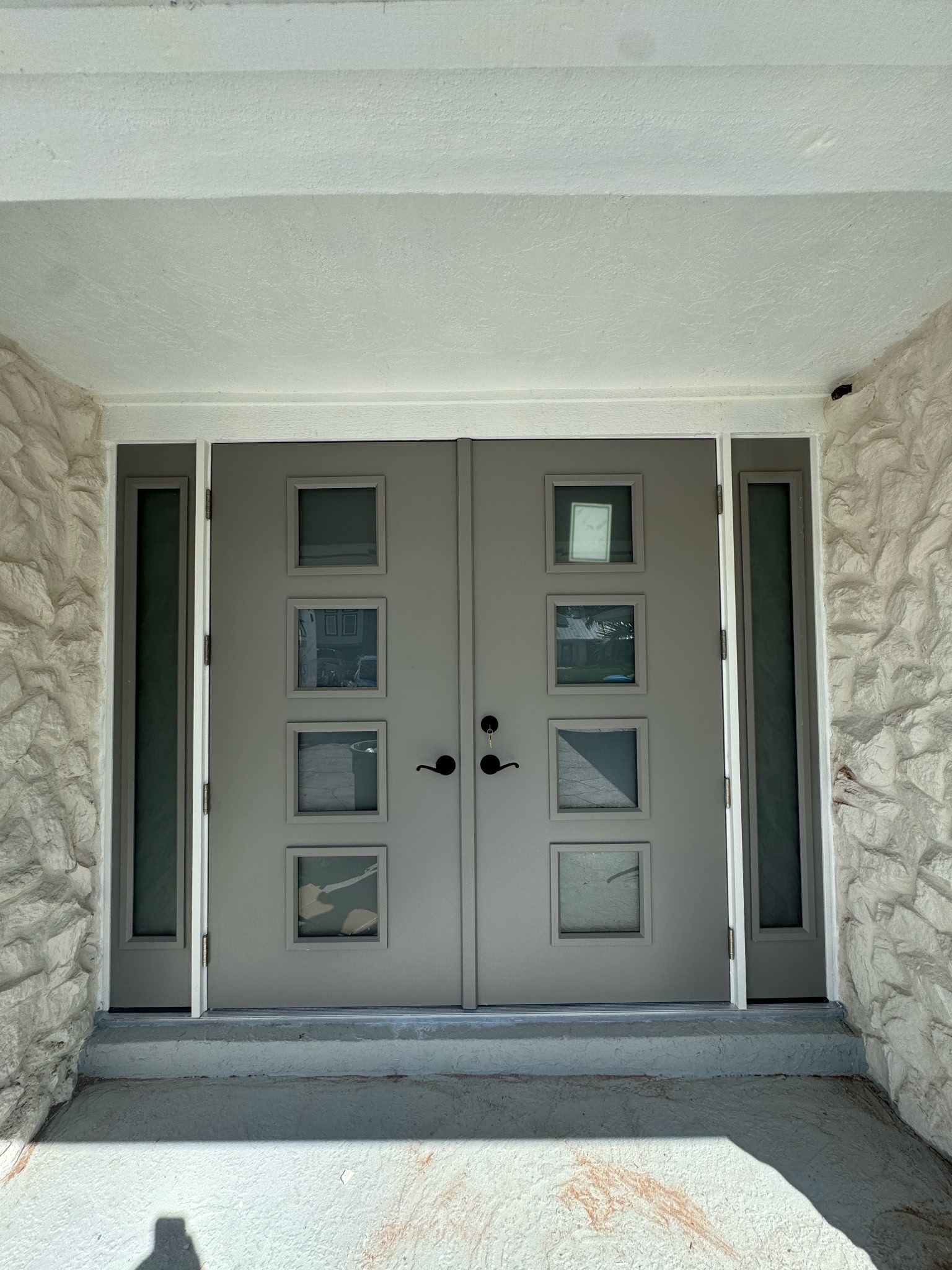 Gray double doors with square windows, flanked by narrow glass panels, set in a stone wall entrance.