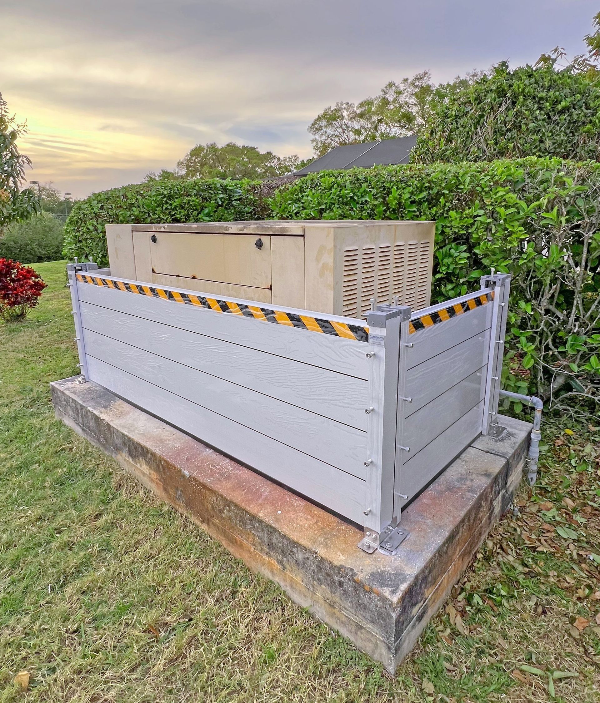 Concrete base with metal barrier, housing outdoor utility equipment. Green yard, foliage, and sky in background.
