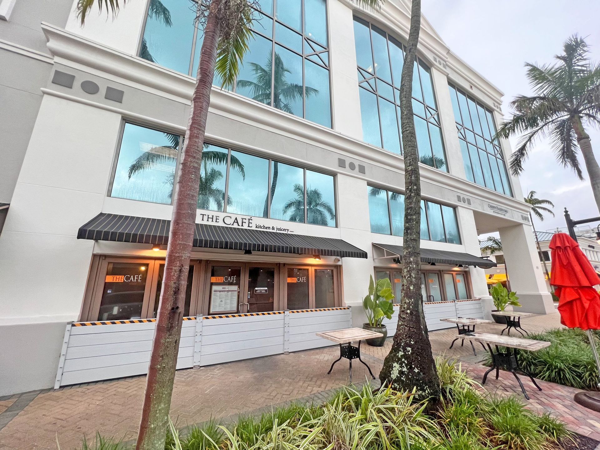 The Cafe, exterior: restaurant with awning, outdoor seating, palm trees, and reflective windows.