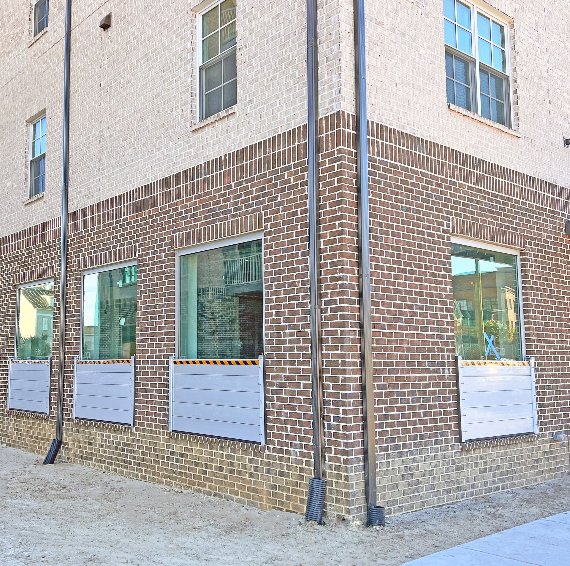 Corner of a brick building with windows and gray panels below. Two tall, dark poles stand by the corner.