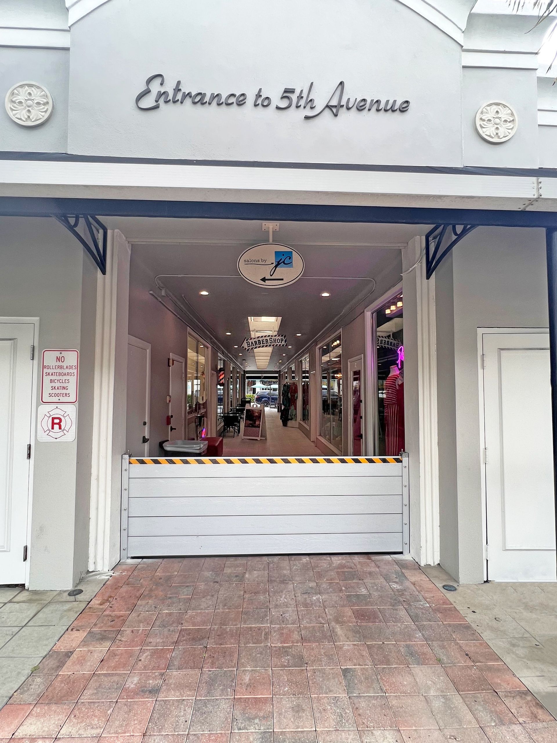 Entrance to 5th Avenue with a flood barrier blocking entry. Gray building exterior and brick walkway.