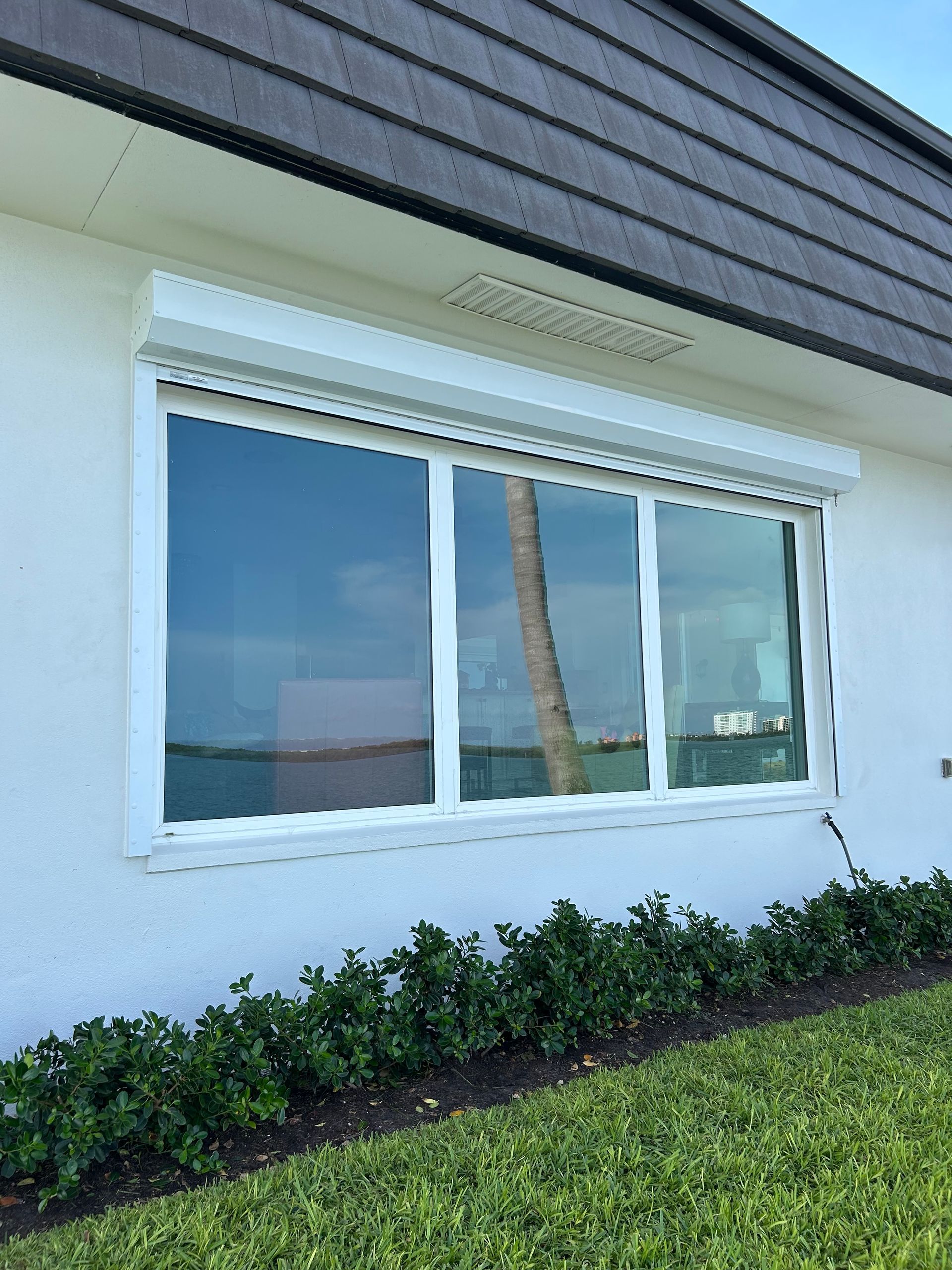White window with roll-down shutter on a white building, overlooking water. Green bushes below.