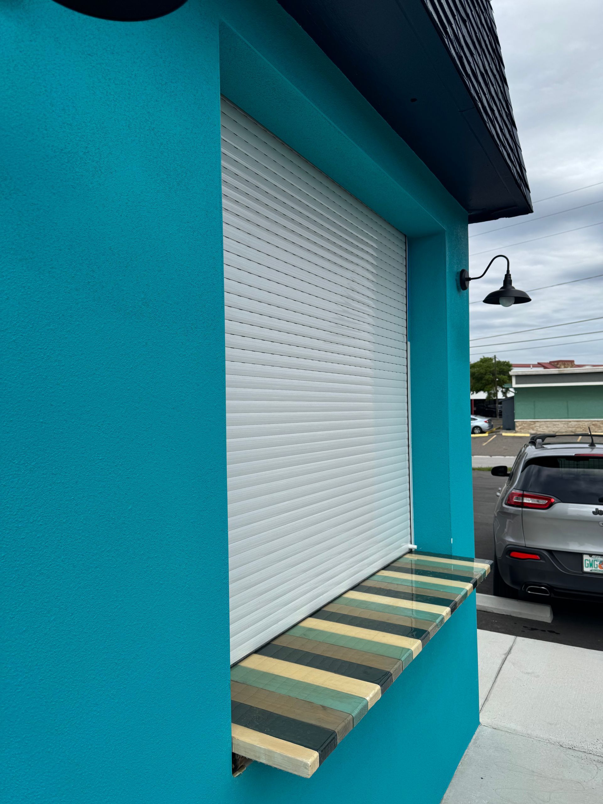 Aqua-colored building with white shuttered window. Striped ledge below window. Black light fixture. Car parked nearby.