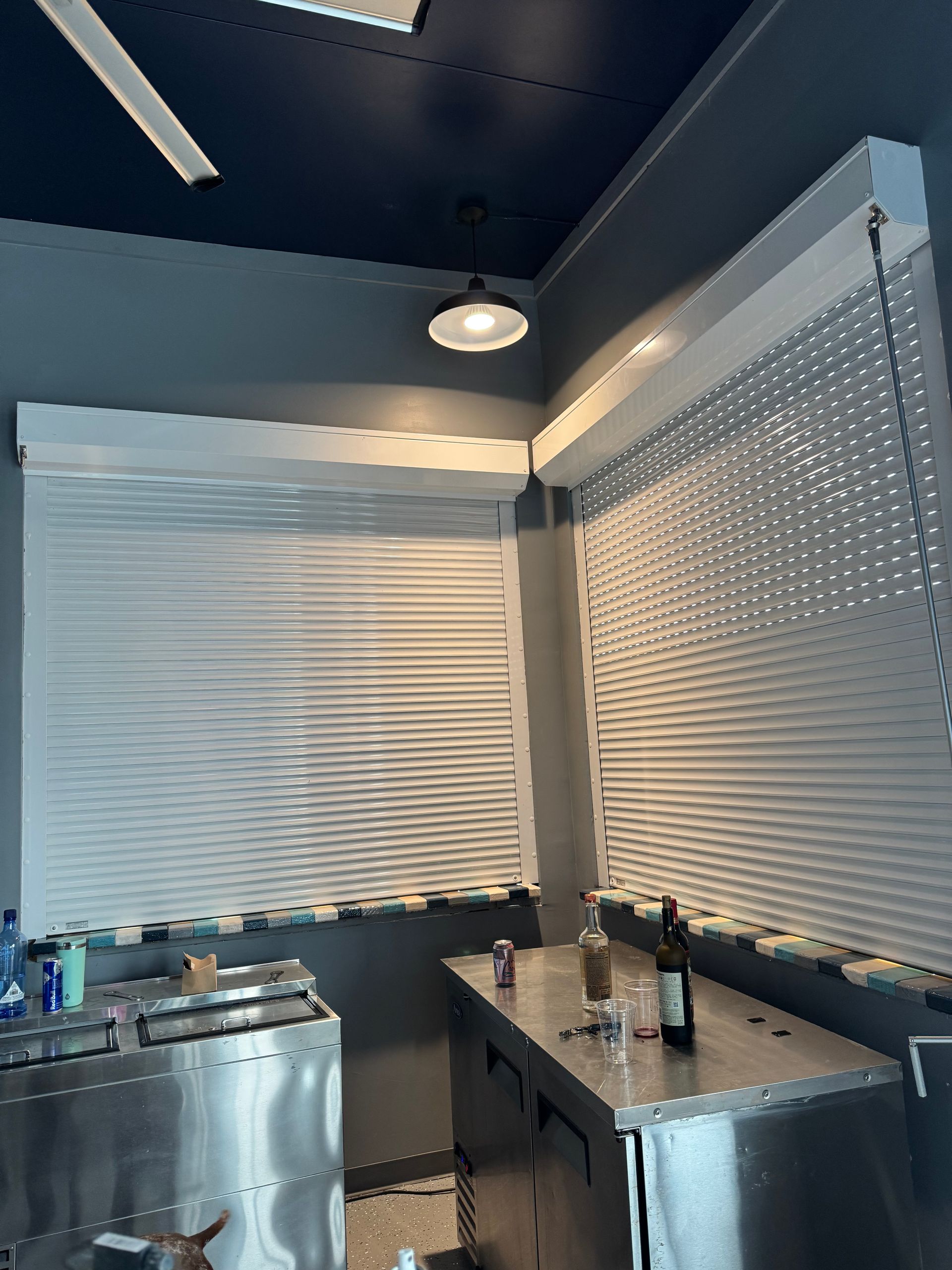 Interior with white blinds, stainless steel counters, overhead lights, dark blue ceiling, and bottles.