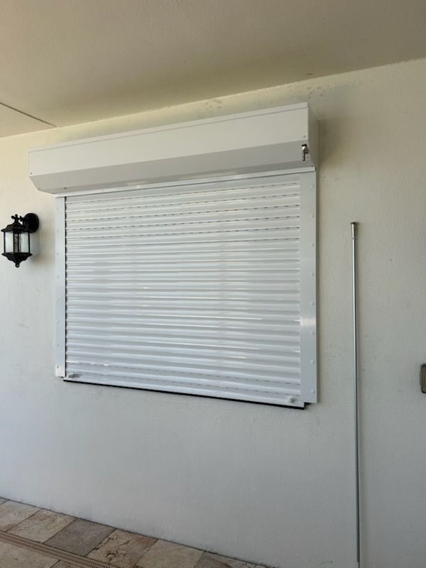 White hurricane shutter over a window on a white exterior wall. A black light fixture is on the wall.