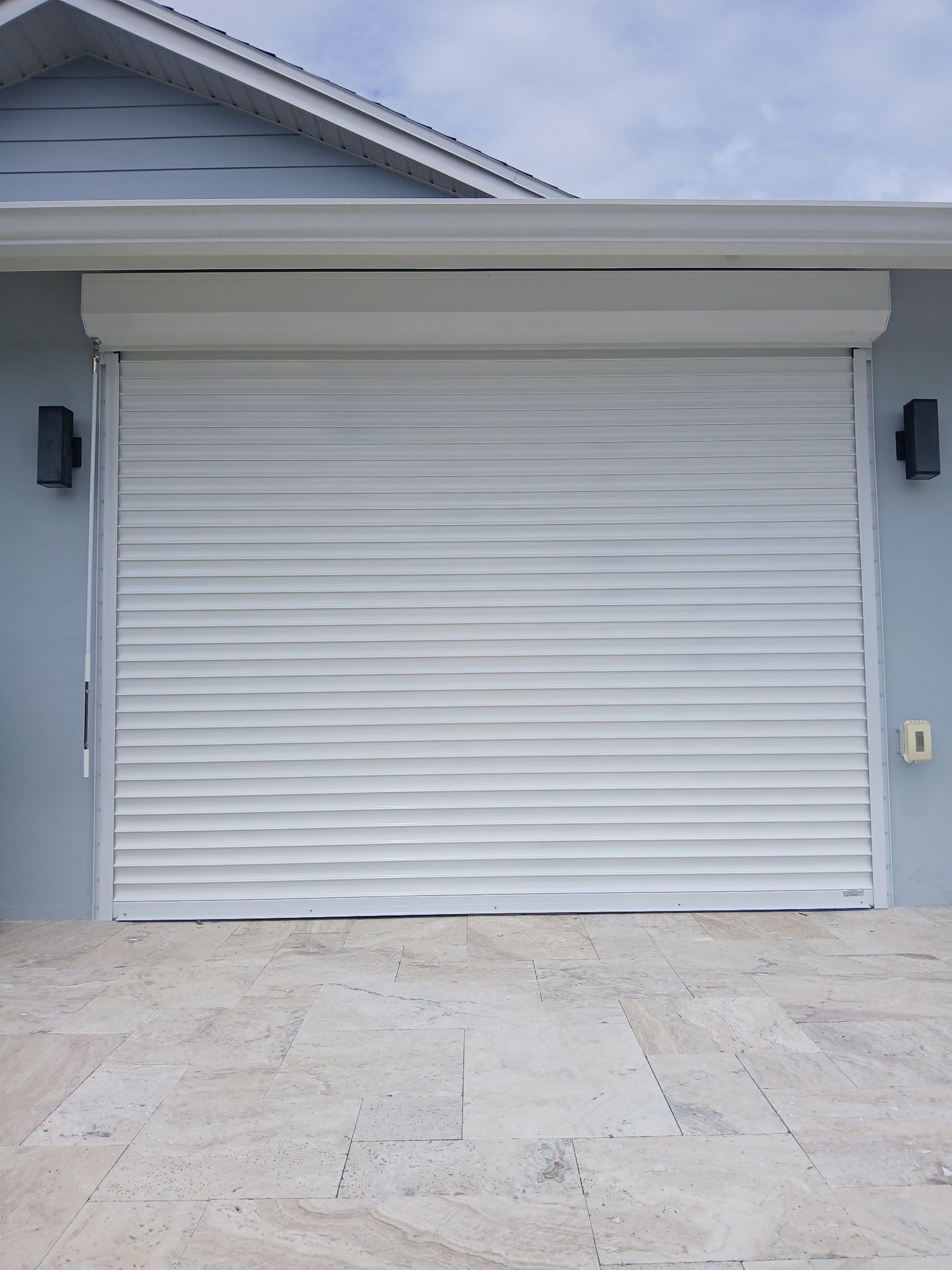 White roll-up security shutter on a light blue building, over a paved driveway, with outdoor lights.
