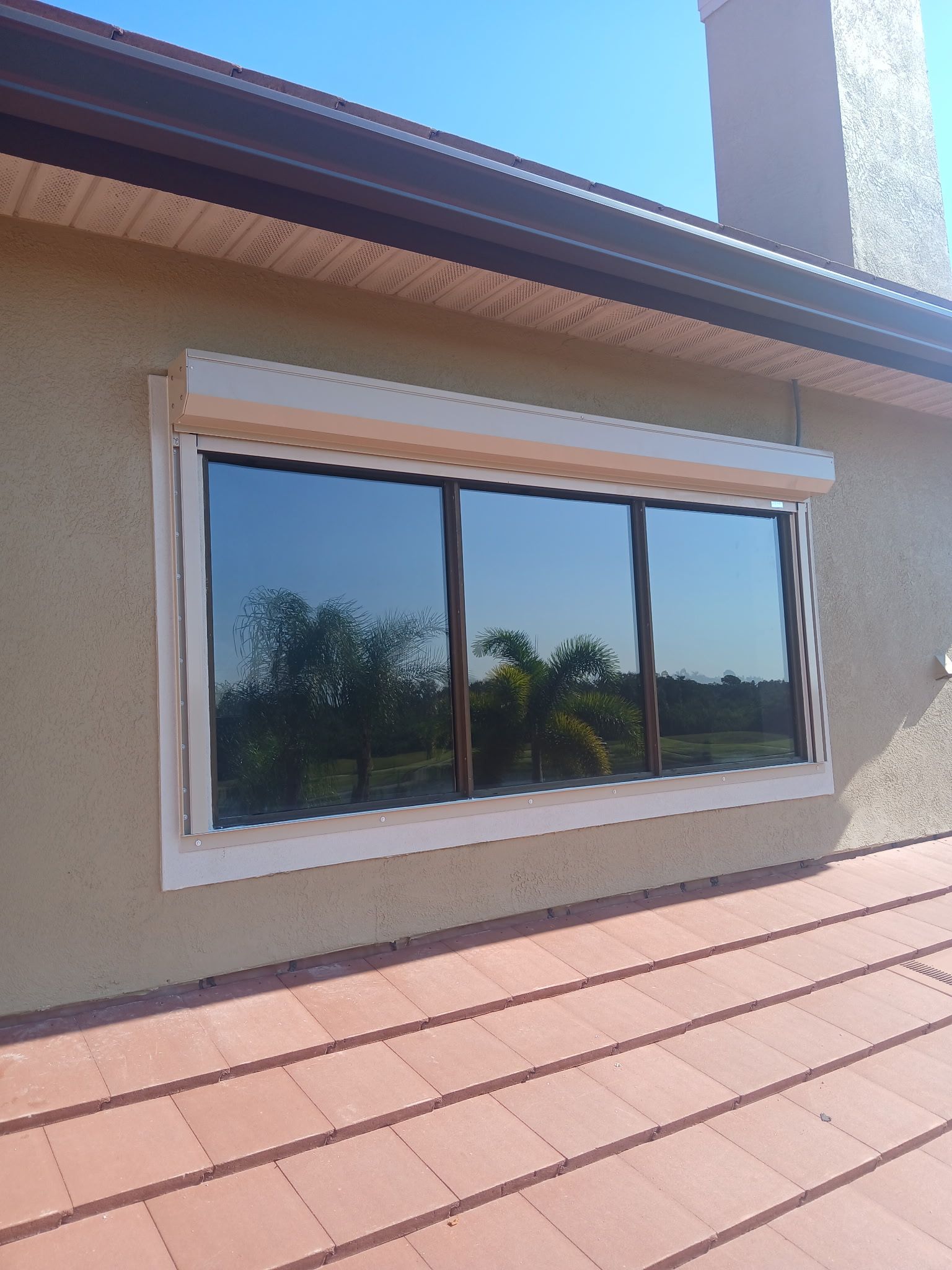 Window with reflective tinted glass, tan stucco wall, brown roof, and a blue sky.