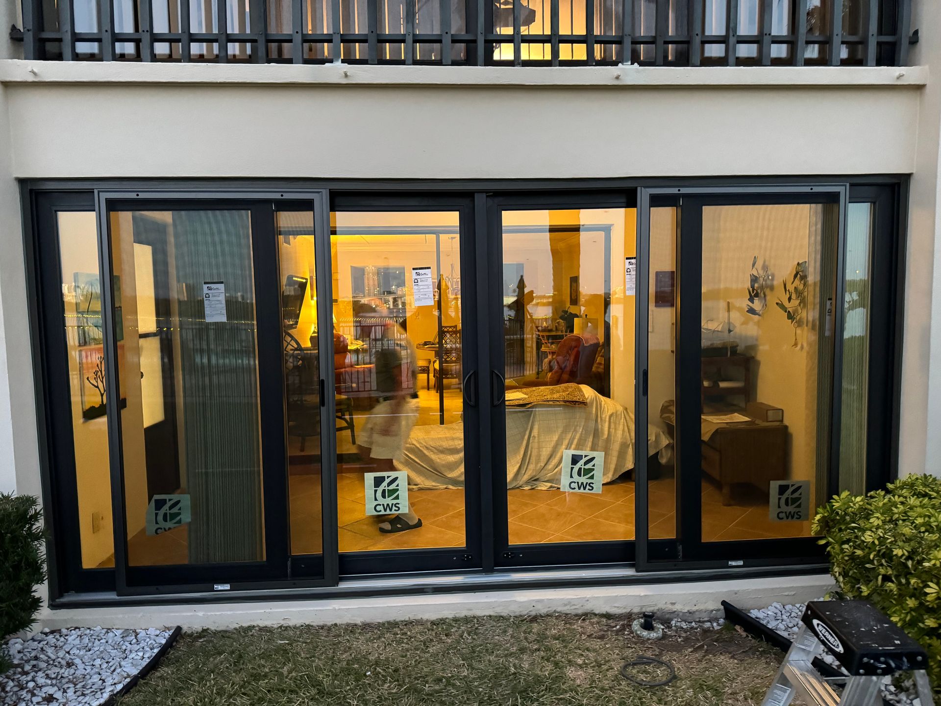 Sliding glass doors with a view of a warmly lit interior. Black frames, beige exterior wall, some greenery.
