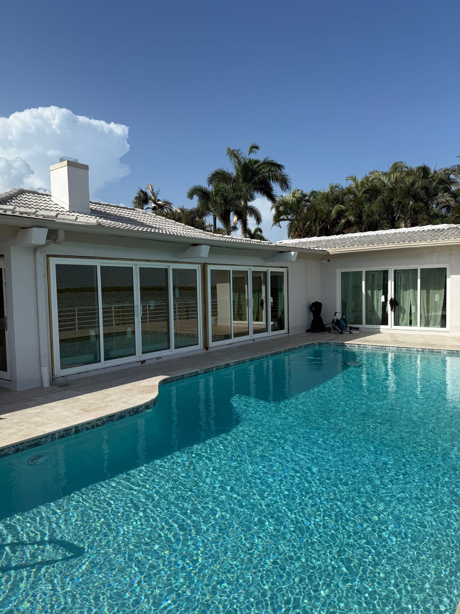 A pool in front of a white house with sliding glass doors, a blue sky overhead, and some trees.