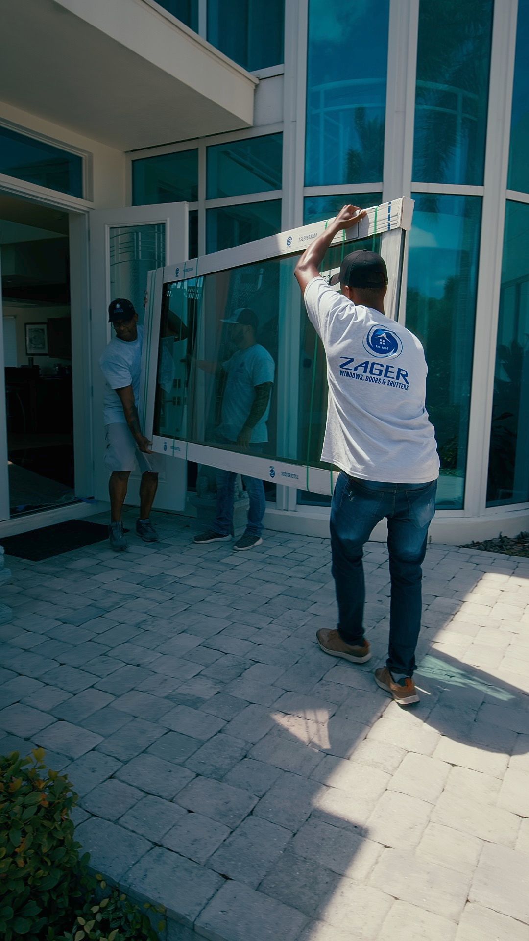 Three people carrying a large window pane towards a modern building with blue glass windows. Sunny outdoor setting.