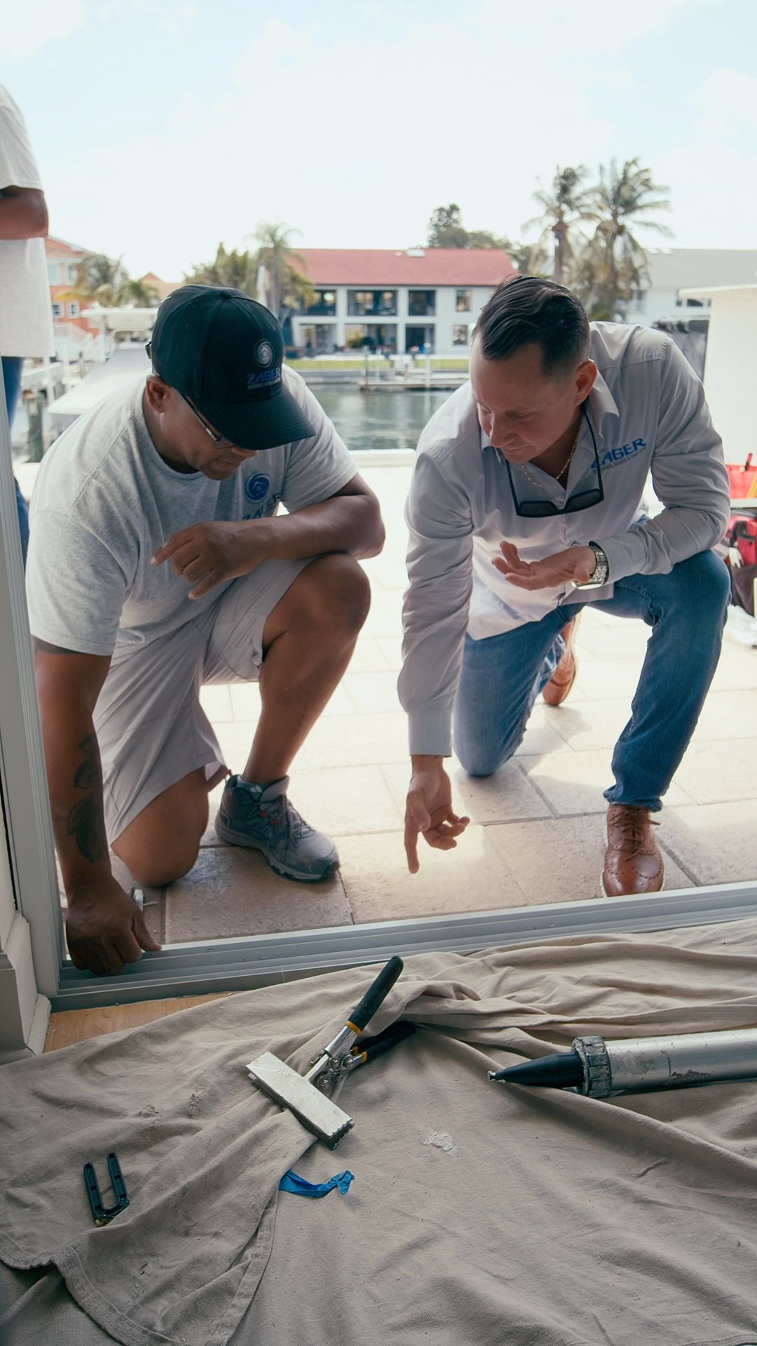 Two men kneel, inspecting a door frame near water. One points; tools and fabric are nearby.