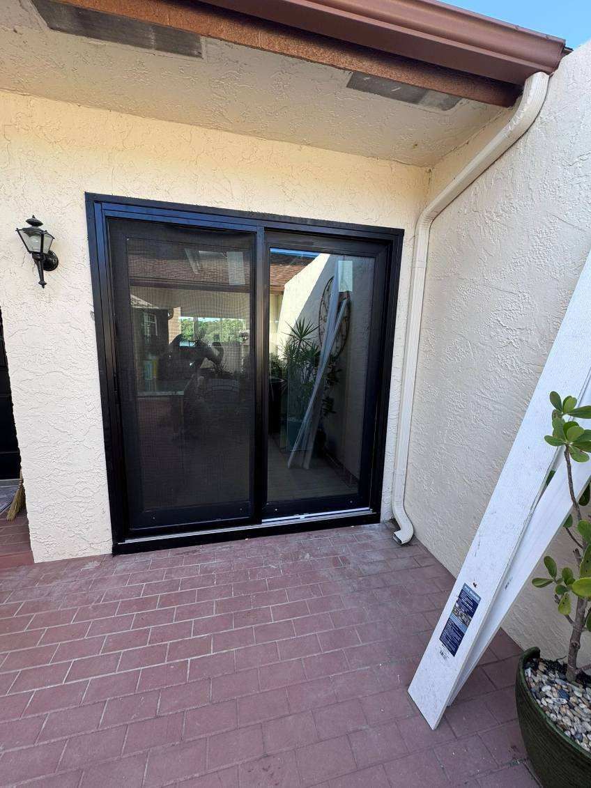 Sliding glass door with black frame on a stucco wall patio. Reflected light, white trim, and a plant.