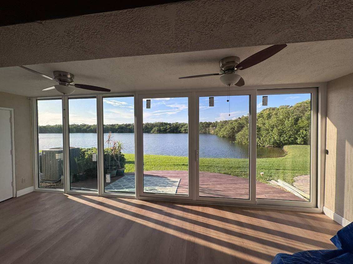 Interior room with large windows overlooking a lake, two ceiling fans, wood floors, and bright sunlight.