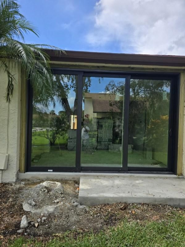 Black-framed sliding glass doors on a concrete patio, reflecting a tree and house, under a brown roof.