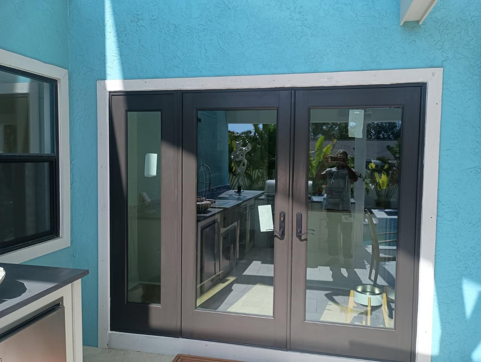 Blue exterior wall with dark framed glass doors and a window, reflecting a patio and outdoor kitchen.