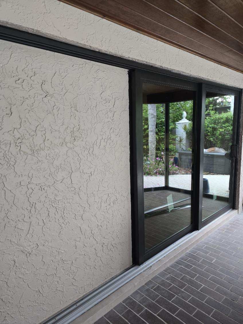 Exterior view of a stucco wall and a black-framed sliding glass door leading to a patio with brick flooring.