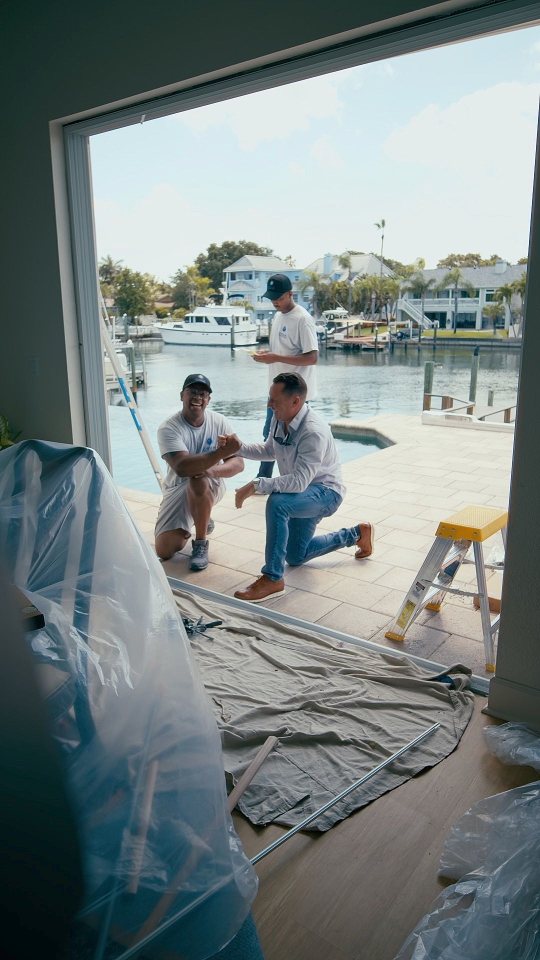 Three men near waterfront. One kneels, pointing. Two others stand. Open doorway frames view of boats.