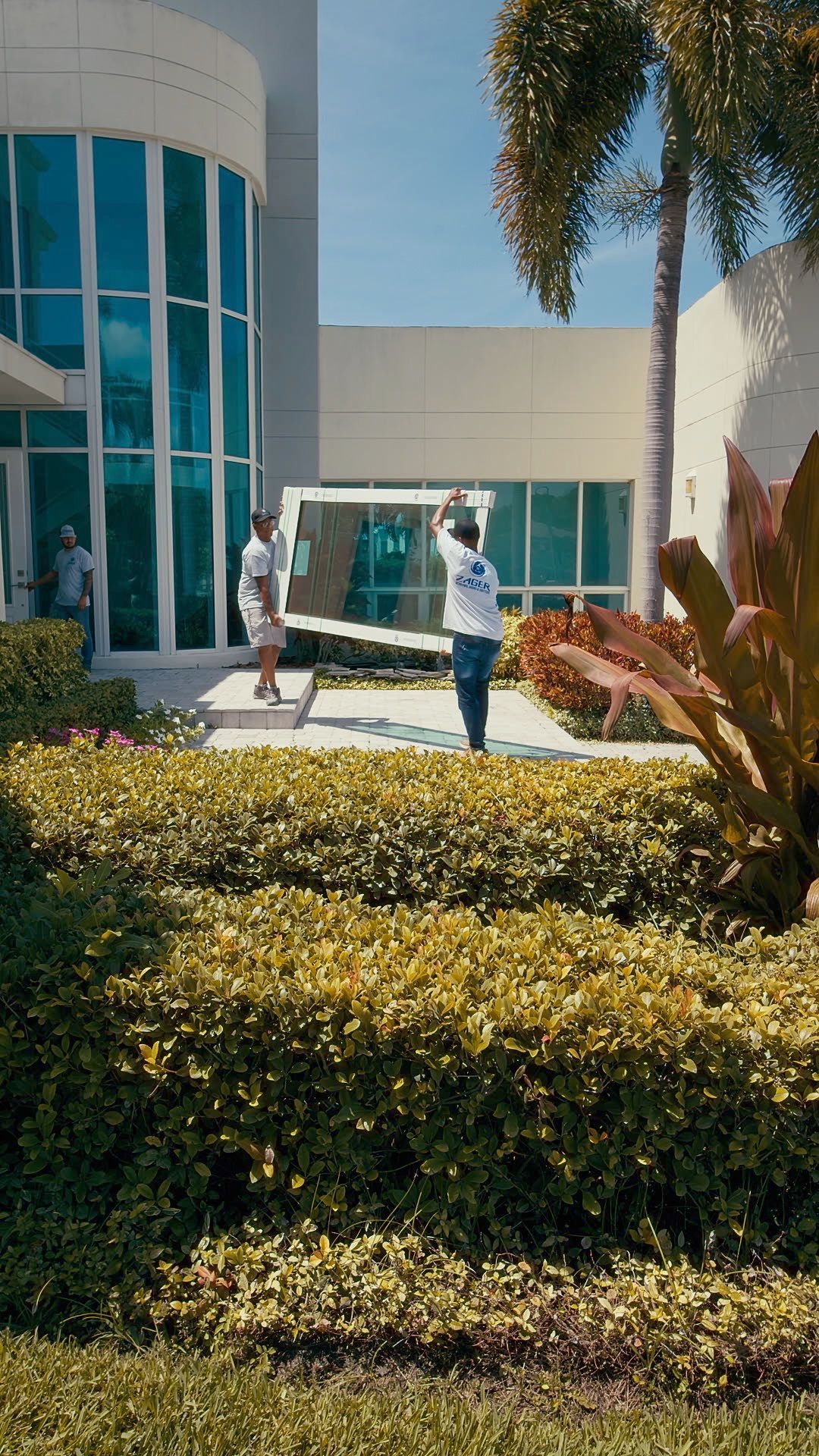 Two people carrying a large pane of glass outside a building with glass windows, sunny day.