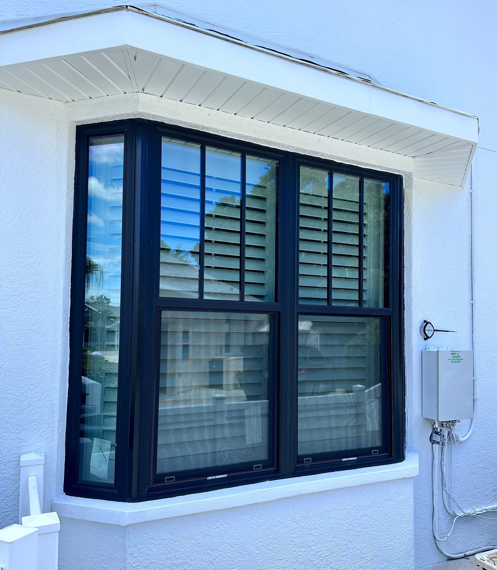 Black framed bay window on white stucco wall, with blinds and reflected sky.