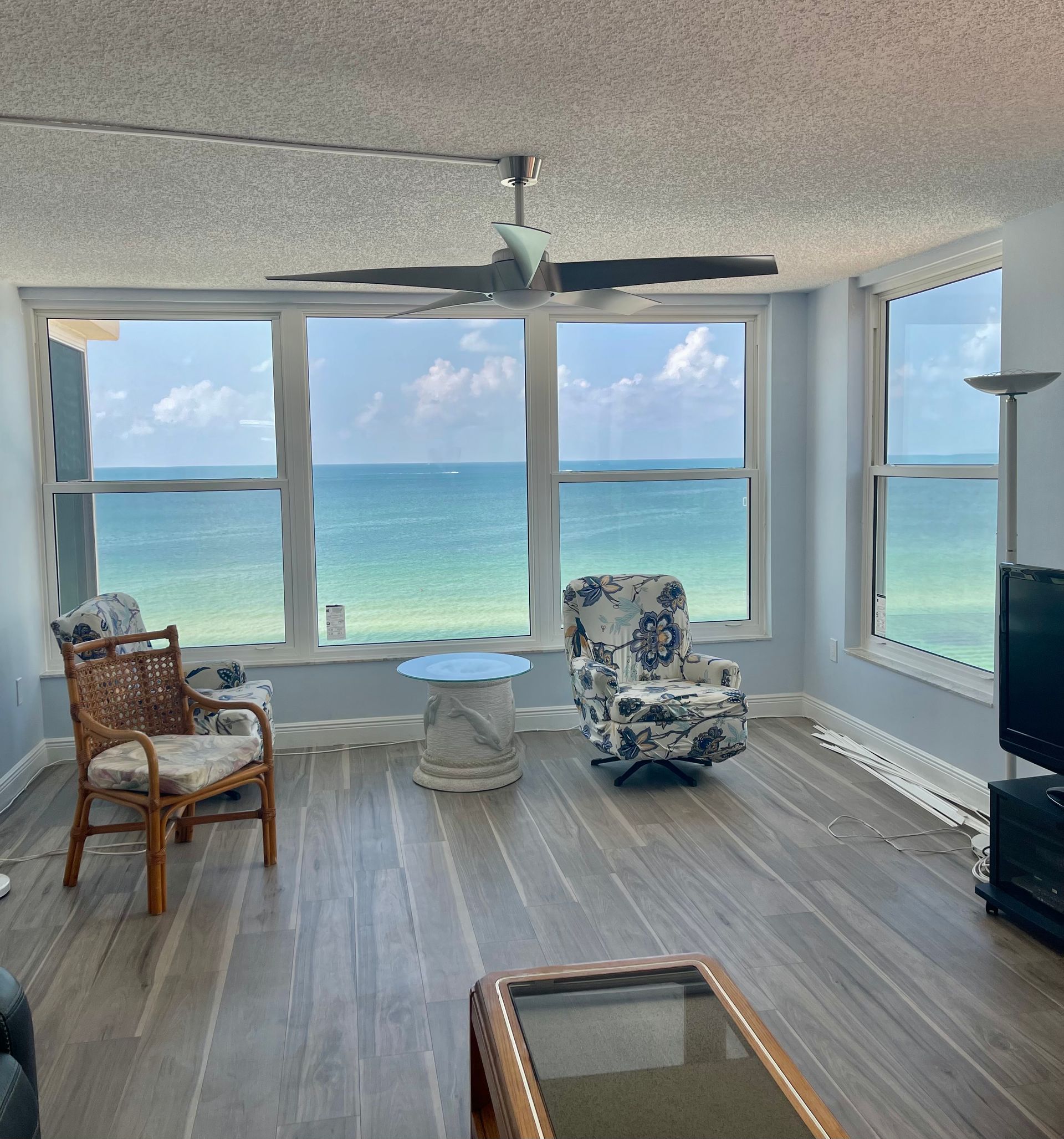Living room with large windows overlooking the ocean. Light blue walls, wood floors, and wicker furniture.