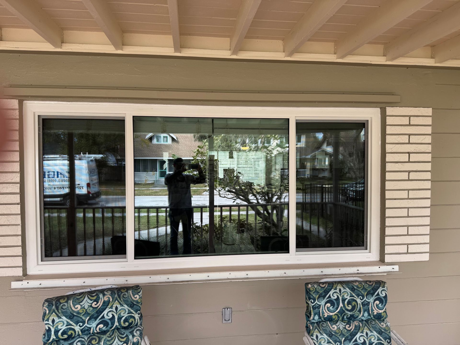 White-framed sliding window reflecting a person, a van, and greenery; on a light-colored building with shutter detail.
