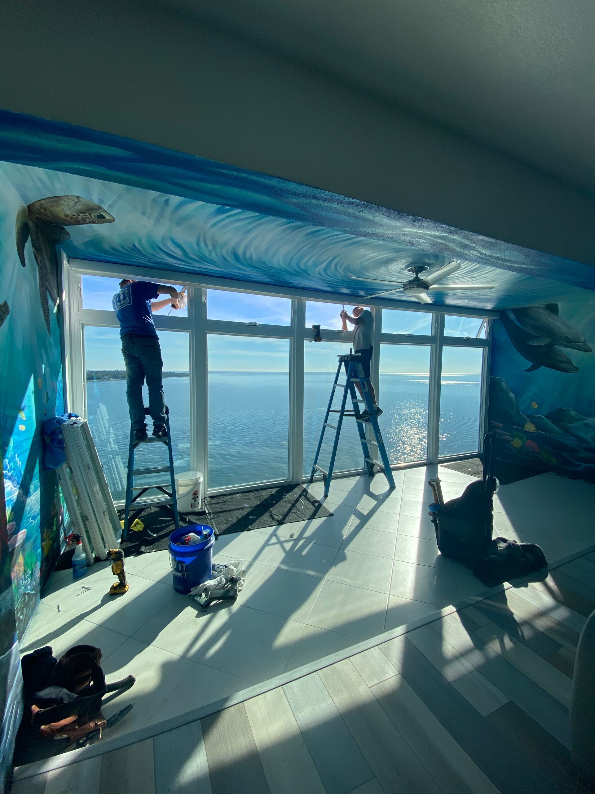 Two people installing window film with ocean view. Ladders, tools, and ocean-themed mural visible.