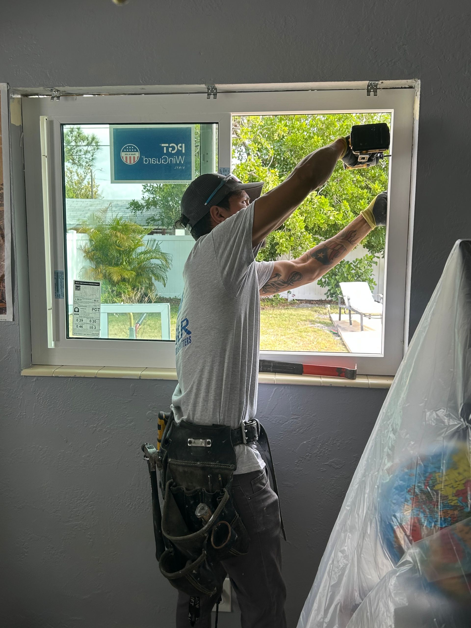 A person installing window; arms up, tools on belt, window frame in place. Green yard visible through the window.