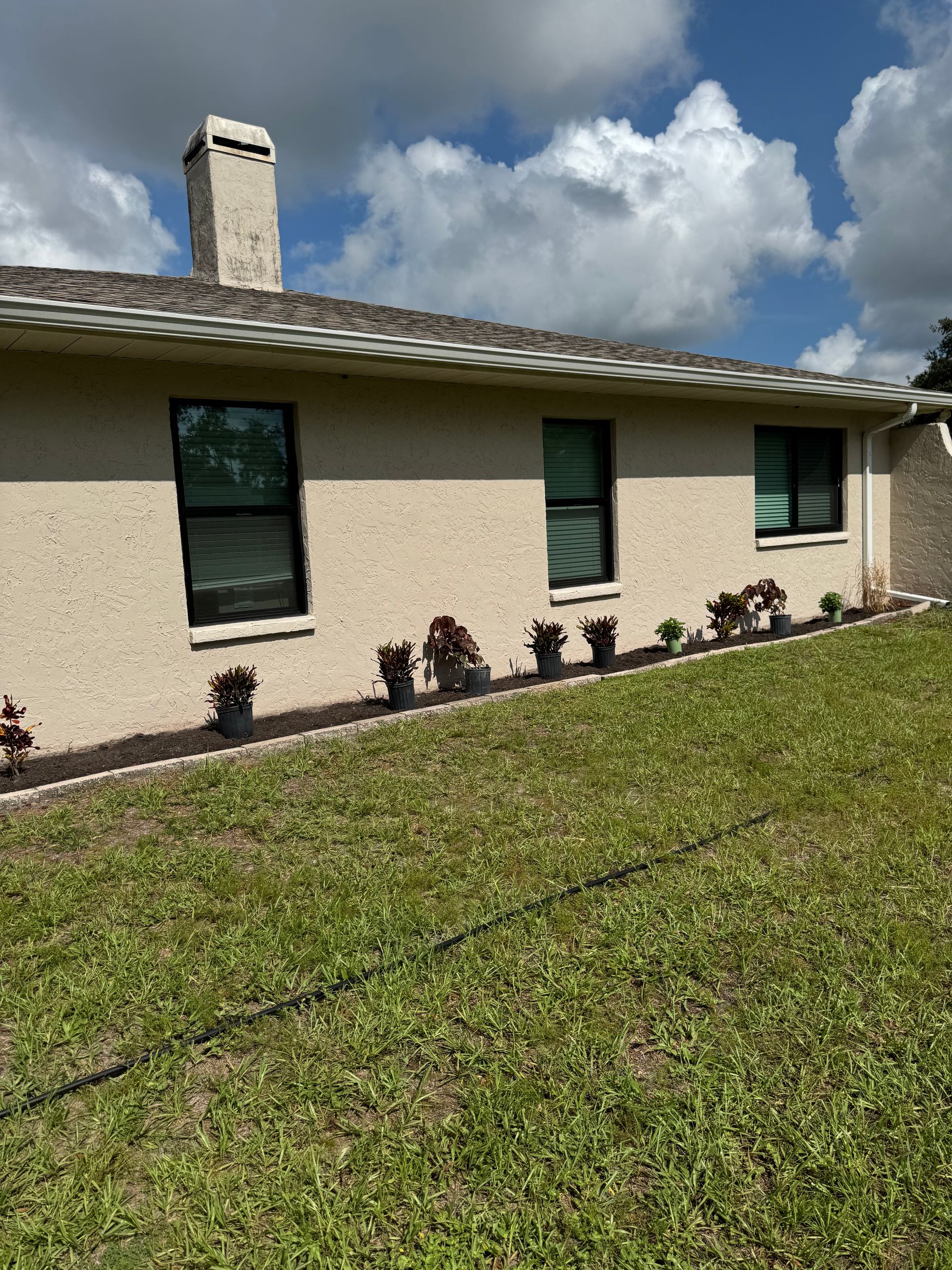 Tan stucco house with black windows, chimney, and a line of potted plants along the foundation, under a partly cloudy sky.