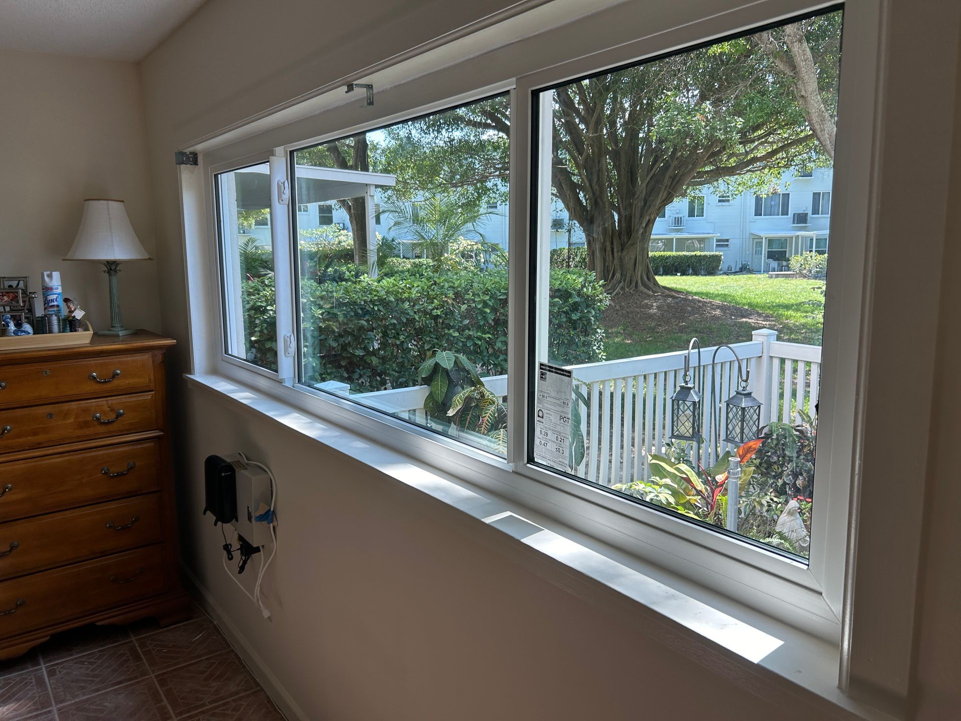 Indoor view of white-framed windows; one is open, revealing a yard with a tree, white fence, and small structure.