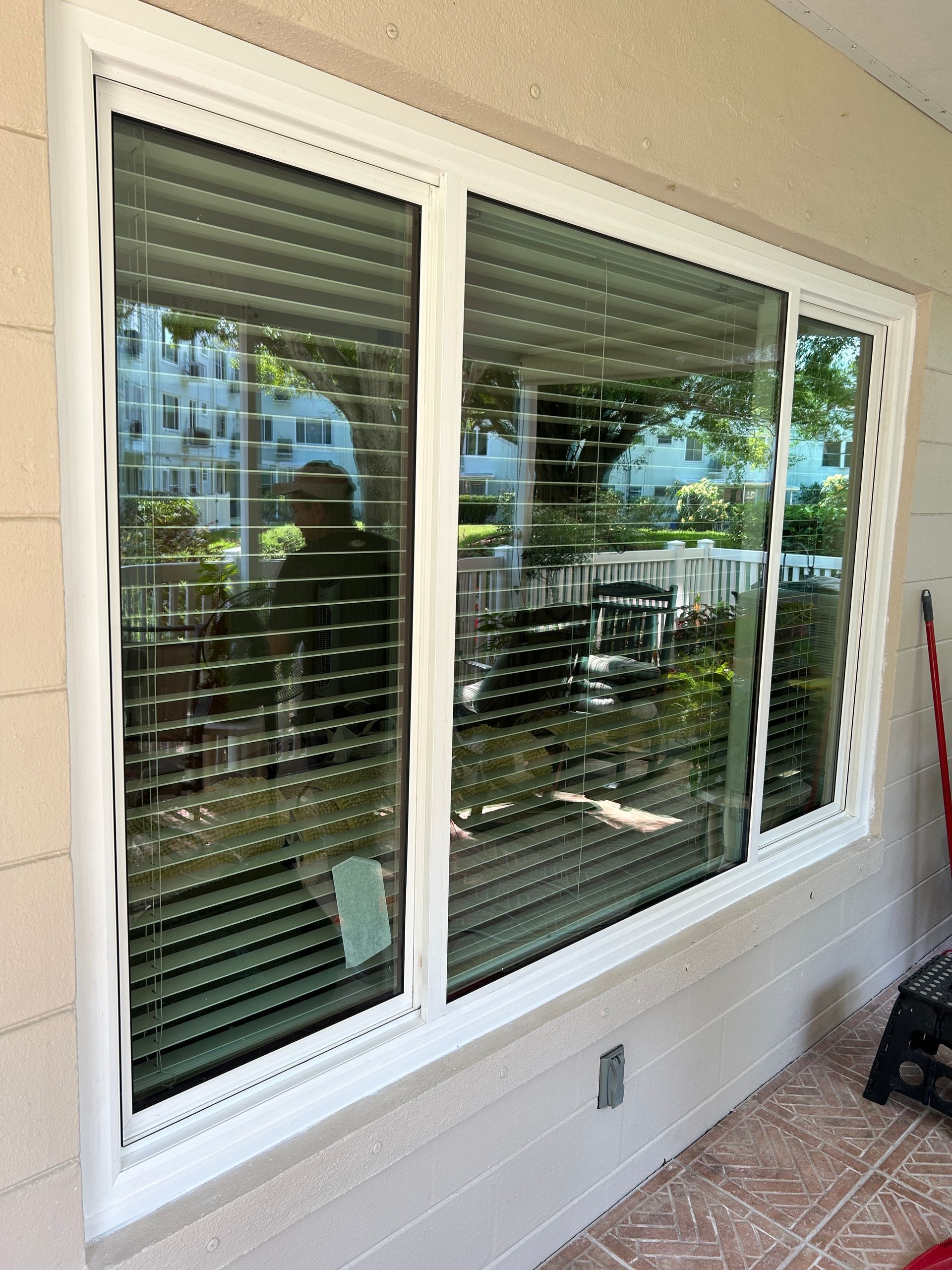 White-framed window with blinds reflecting outdoor scene. Window is set in a light beige exterior wall, over a brick patio.