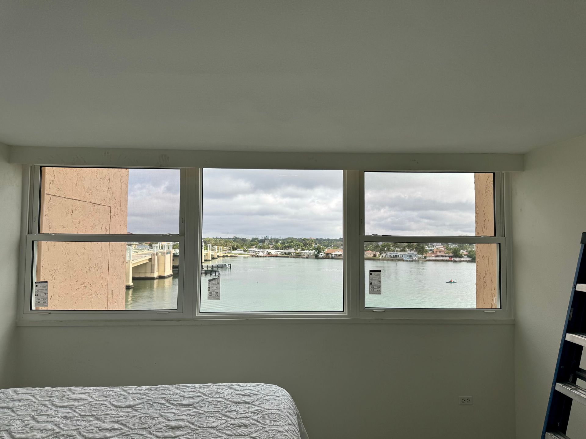 A wide window overlooking a body of water with a bridge and cloudy sky.