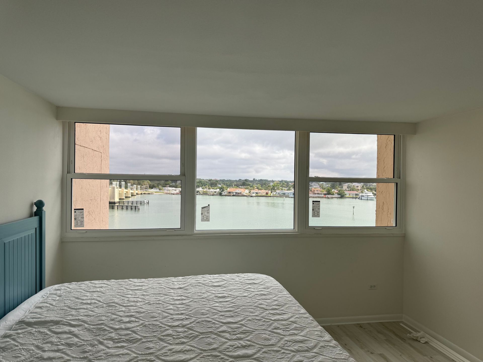 Bedroom with large windows overlooking a waterfront scene; bed with blue headboard is in the foreground.