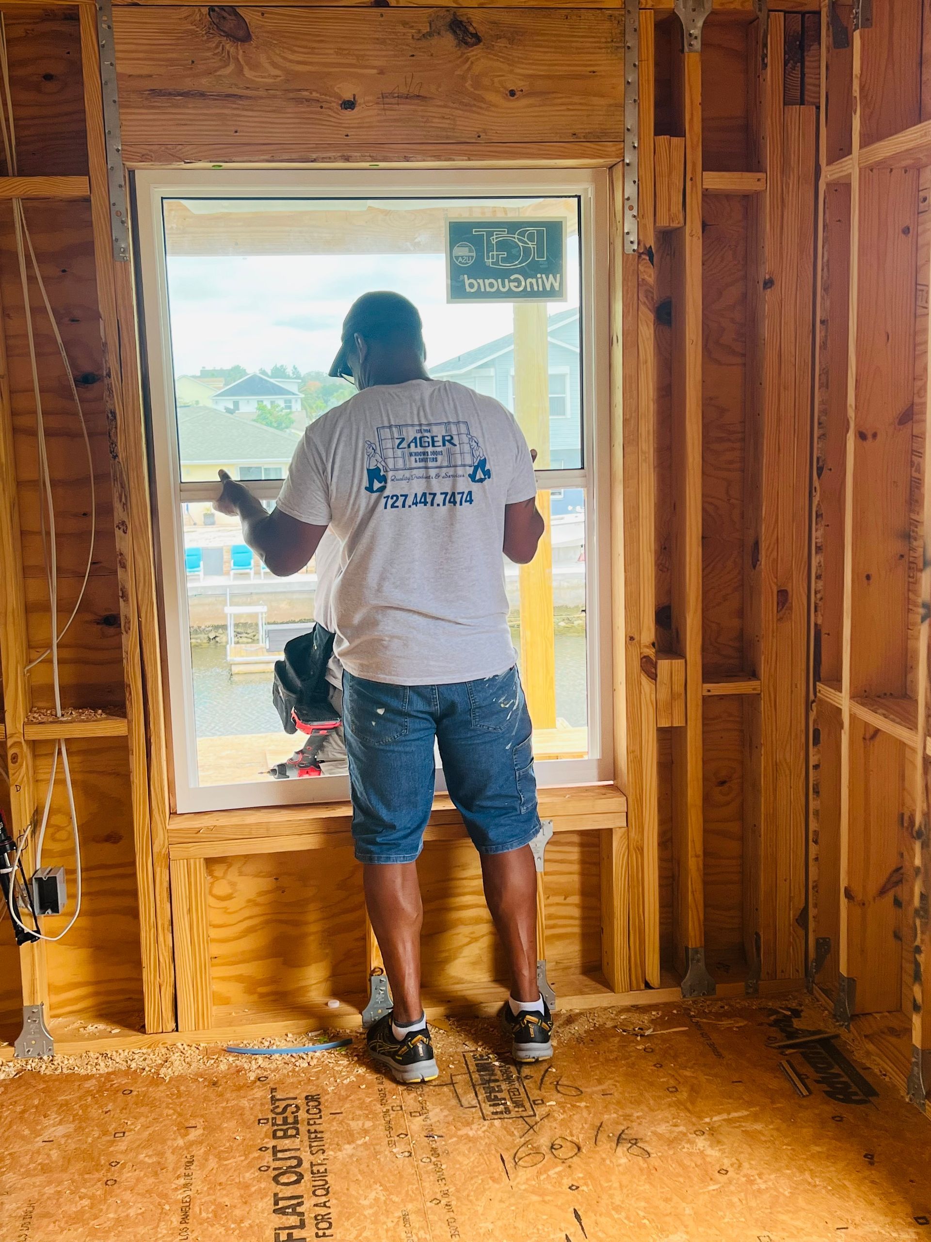 Man installing a window in a new construction. He wears a hat, shorts, and a t-shirt.