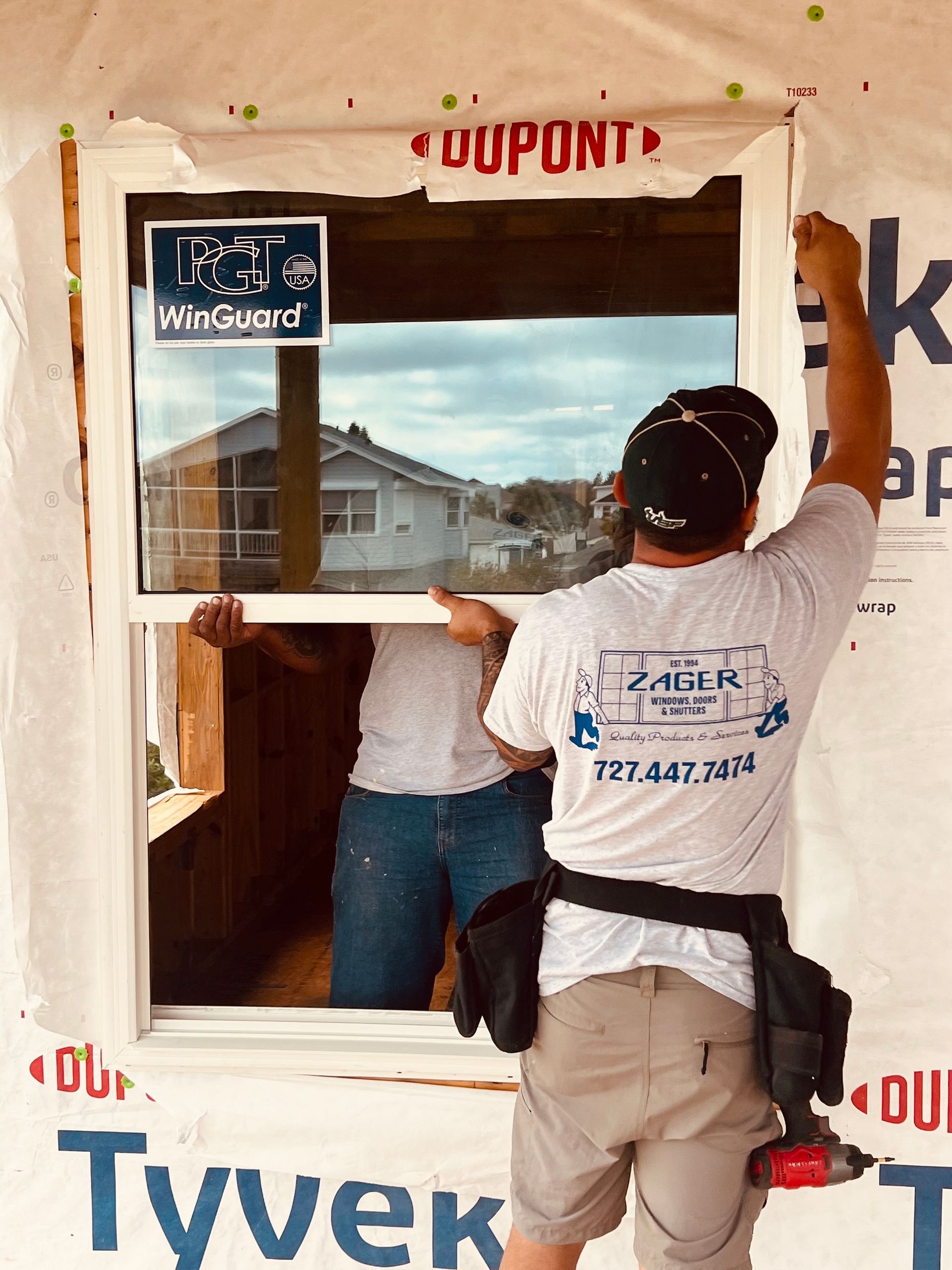 Two people installing a window in a house under construction. One person is wearing a white shirt and holding the window.