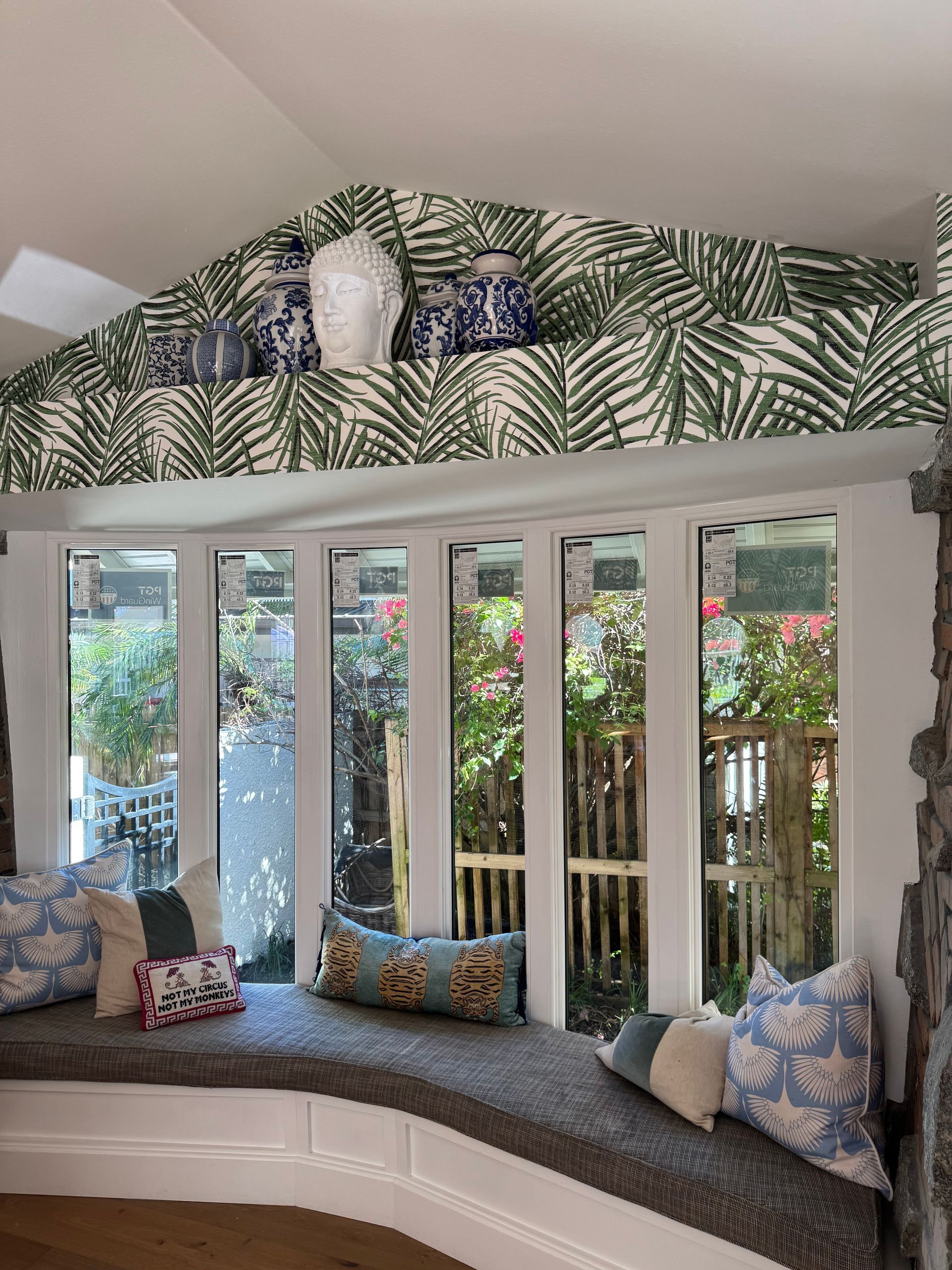 Bay window with cushion seating, decorated with patterned pillows. Palm frond wallpaper and white trim.