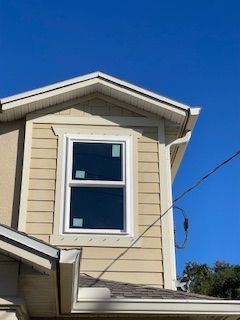 Exterior view of a house with a window on the second story, surrounded by beige siding and white trim against a blue sky.